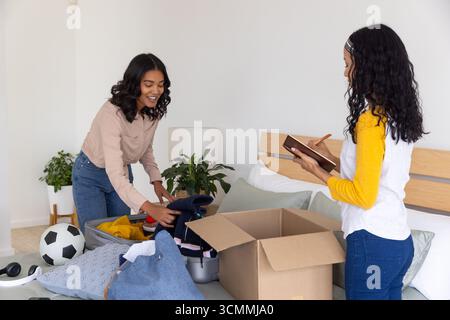 Mamma e figlia adolescente diverse che ordinano i vestiti a casa, appoggiandosi sulla valigia Foto Stock