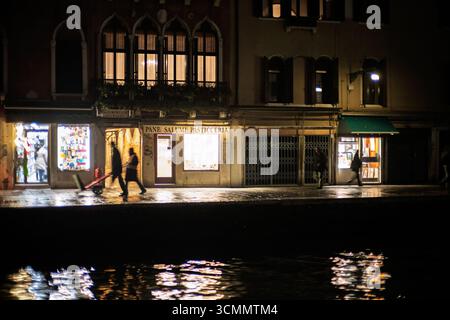 I riflessi danzano sull'acqua mentre i passanti sfocati passeggiano lungo il Canale di Cannaregio, illuminati da calde luci del negozio nel cuore di Venezia. Foto Stock