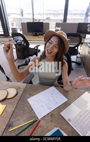 Donna ispanica che indossa un cappello seduto alla scrivania in ufficio mentre gestiva gli schizzi con matite colorate Foto Stock
