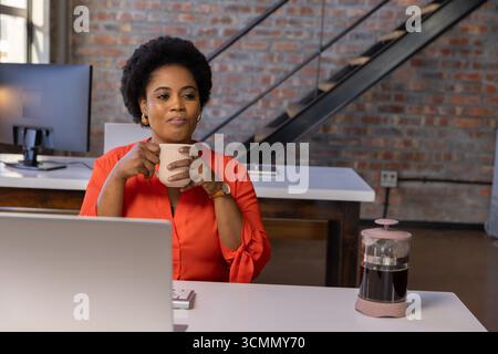 Donna afroamericana di medie dimensioni che tiene in mano una tazza da caffè e lavora su un computer portatile alla scrivania in ufficio Foto Stock