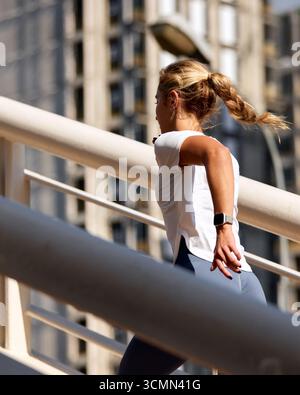 Una giovane donna che indossa abbigliamento sportivo corre lungo un ponte in un ambiente urbano. La scena cattura la sua dedizione al fitness e a uno stile di vita attivo, illu Foto Stock