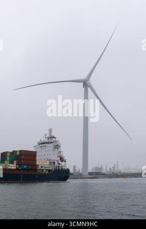 La nave portacontainer EA Gannet è ormeggiata in una giornata nebbiosa nel porto di Rotterdam, Paesi Bassi, con una turbina eolica che si innalza sullo sfondo sopra il lungomare industriale e acque calme. Foto Stock
