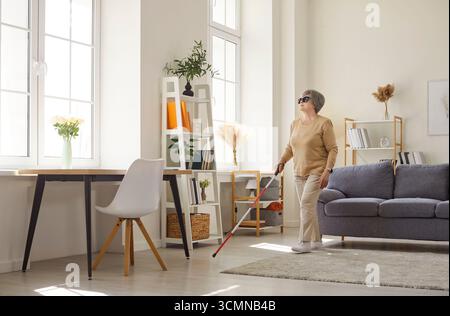 Anziani anziani non vedenti disabili in abiti casual con canna che cammina nel soggiorno di casa. Foto Stock