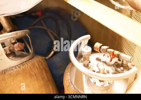 Primo piano che mostra due bombole di gas in metallo con valvole e regolatori di pressione collegati, posizionate all'interno del vano tecnico o industriale, tubi flessibili e raccordi visibili sullo sfondo Foto Stock