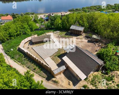 Vista aerea ad alto angolo di un sito storico con capanne in legno ricostruite e strutture in legno, con il fiume Oka e la sua riva sabbiosa sullo sfondo Foto Stock