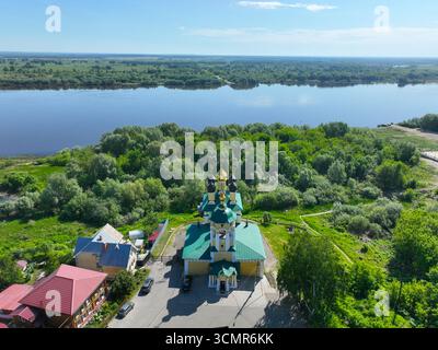 Una vista aerea di una vivace chiesa gialla con tetti verdi e cupole dorate, con una lussureggiante foresta verde e l'ampio fiume Oka sullo sfondo. Foto Stock