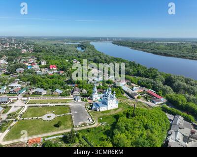 Un'ampia vista aerea che mostra una chiesa bianca con cupole blu in primo piano, con il tortuoso fiume Oka e il ponte Murom sullo sfondo. Foto Stock