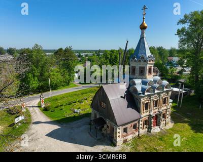 Una vista aerea di una cappella in mattoni ornata con un tetto blu e una cupola dorata, annidata in un piccolo villaggio con una strada e dintorni verdi. Foto Stock