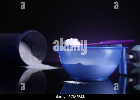 Prodotto cosmetico biondo per la cura dei capelli. Polvere nel recipiente, nel recipiente e nella spatola sulla superficie a specchio nera su sfondo viola scuro, primo piano Foto Stock