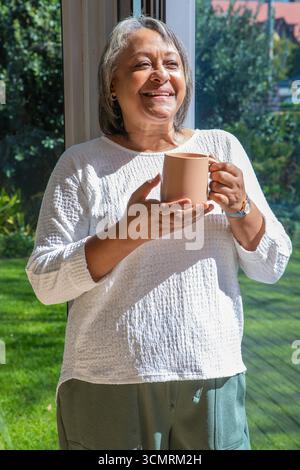 Donna afroamericana anziana che tiene la tazza da caffè accanto a una porta di vetro e guarda il giardino Foto Stock