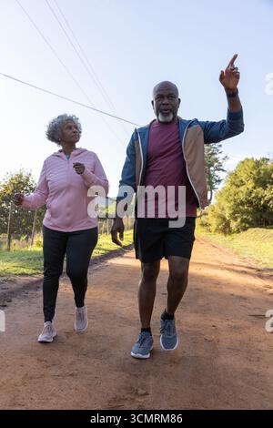 Coppia senior afroamericana che cammina su una strada sterrata che punta verso l'alto in felpa con cappuccio sotto le linee elettriche Foto Stock