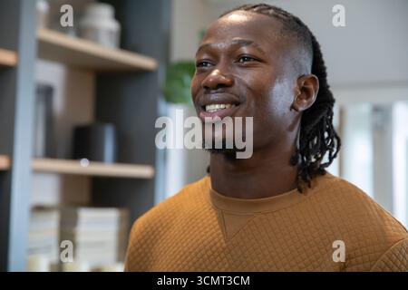 Uomo afroamericano sorridente in felpa marrone che guarda verso l'alto in ufficio con scaffale Foto Stock