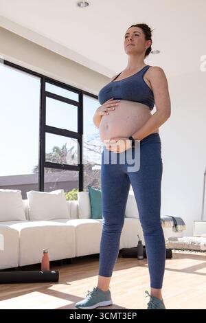 Donna incinta in piedi in un luminoso soggiorno che culla la pancia vicino a un divano a sezione bianca e al tappetino per yoga Foto Stock