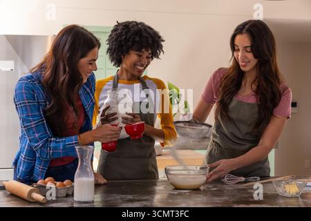 Diverse amiche che misurano la farina, versano la pastella nella ciotola di vetro dell'isola della cucina di casa Foto Stock