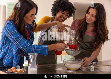 Diverse amiche che mescolano la pastella all'isola della cucina con vasetti di farina e misurini Foto Stock