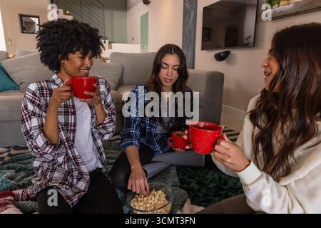 Diverse amiche con tazze rosse e popcorn su una lussuosa coperta in soggiorno Foto Stock