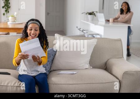 Mamma e figlia adolescente diverse che usano il laptop e leggono una lettera sul divano a casa, copiano lo spazio Foto Stock