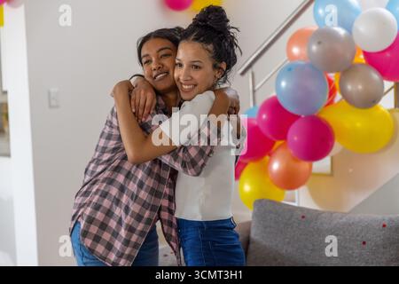 Mamma e figlia adolescente diverse che si abbracciano nel soggiorno vicino alla scala con palloncini Foto Stock