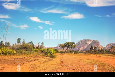 Strada rossa di polvere e fango, piccoli cespugli e alberi sui lati e sulle montagne - il massiccio di Andringitra - sullo sfondo, tipico scenario vicino ad Ambalavao Foto Stock