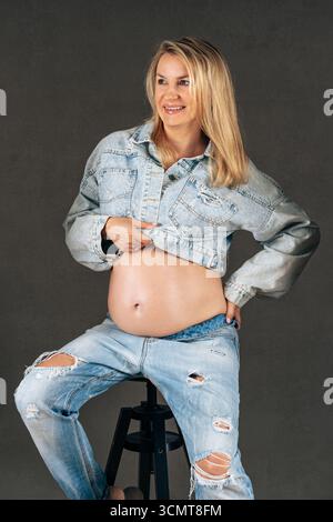 Moderno ritratto in studio di una donna incinta in jeans strappati e giacca in denim. Concetto di stile di vita in gravidanza, anticipazione e maternità Foto Stock
