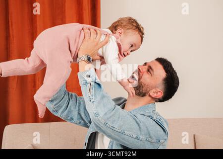 Un padre gioioso si impegna in un'interazione giocosa mentre solleva il suo bambino sorridente, mostrando un momento commovente pieno di risate e amore Foto Stock