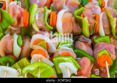Vista ravvicinata degli spiedini di pollo crudi con peperoni verdi, cipolle e carote su bastoncini di legno, pronti per la cottura alla griglia o in estate. Foto Stock