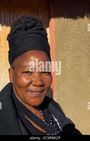 donna africana single del villaggio con trecce di dreadlock, seduta davanti alla casa , nel cortile, con un sorriso Foto Stock