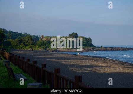 La sabbia nera della spiaggia di Hahyo Soesokkak si estende verso un punto roccioso e un promontorio ricoperto di palme, mentre piccole onde lambiscono le pietre alla bocca del so Foto Stock