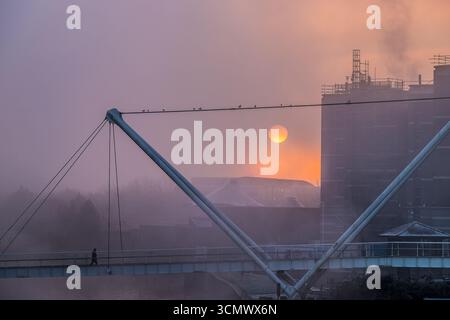 Suggestiva scena mattutina a Leeds che mostra un ponte pedonale sul fiume Aire con il sole che sorge attraverso la fitta nebbia Foto Stock
