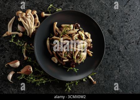 Shimeji fritti con timo e aglio su un piatto nero. Funghi freschi e preparati con ingredienti. Cucina tradizionale giapponese. Vista dall'alto. Foto Stock
