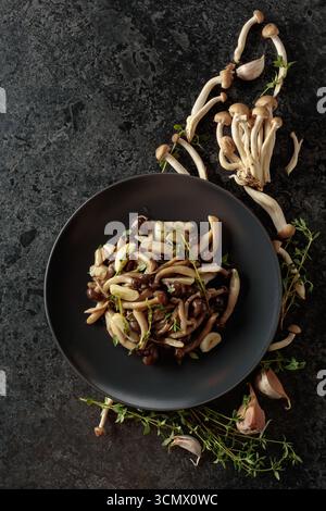 Shimeji fritti con timo e aglio su un piatto nero. Funghi freschi e preparati con ingredienti. Cucina tradizionale giapponese. Vista dall'alto. Foto Stock