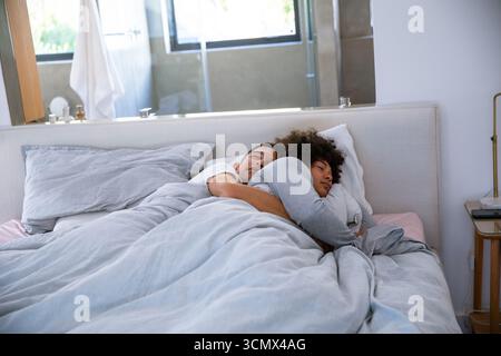 Due uomini che si coccolano sul letto sotto un piumone grigio chiaro nella camera da letto principale vicino allo smartphone sul comodino Foto Stock
