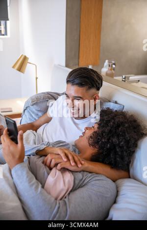 Due uomini sdraiati sul letto ridono mentre navigano sullo smartphone in camera da letto con lavandino e lampada da comodino Foto Stock