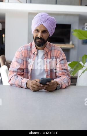 Uomo indiano seduto al tavolo in camera che tocca lo smartphone accanto alla televisione a schermo piatto Foto Stock