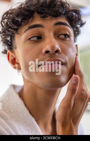 Uomo che tocca la guancia durante la routine di dermocosmesi in bagno vicino alla finestra e indossa un accappatoio di spugna Foto Stock