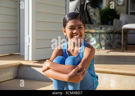 Bambina asiatica seduta abbracciando le ginocchia sorridendo sui gradini del portico di legno con sedia da patio, pianta in vaso Foto Stock