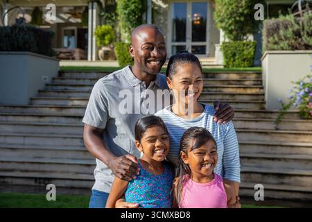 Famiglia variegata in piedi sul prato di fronte a scalini di pietra e siepi rifilate all'ingresso della casa Foto Stock