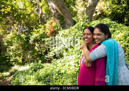 Madre e figlia indiane che abbracciano e sorridono in un lussureggiante giardino con kurta colorati e spazio copia Foto Stock