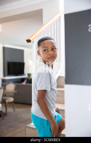 Ragazzo afroamericano in età scolare scalata a casa da banister e sguardo lasciato alla TV Foto Stock