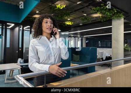 Donna afroamericana che parla sullo smartphone accanto a una ringhiera in legno in un ufficio open space Foto Stock