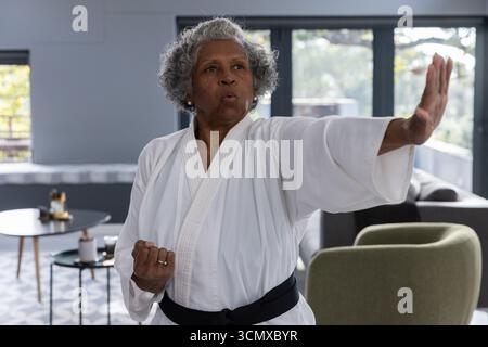 Donna afroamericana anziana che pratica il blocco di karate indossando gi, cintura nera in soggiorno Foto Stock