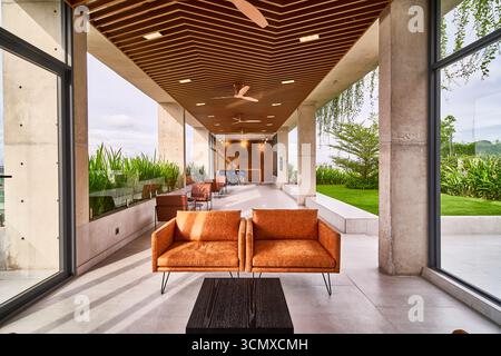 Lussuoso corridoio aperto con divani arancioni, porte in vetro, soffitto in legno e lussureggianti viste esterne, che mettono in risalto il design architettonico e l'eleganza moderni. Foto Stock