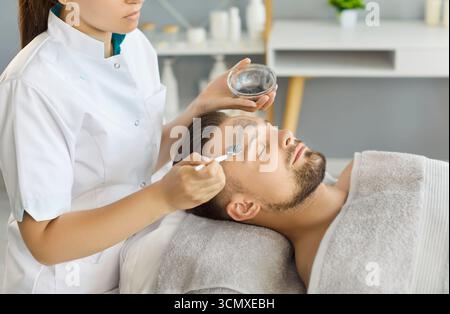 Cosmetologo che tiene la ciotola e la spazzola, applicando la maschera facciale a un giovane cliente maschio Foto Stock