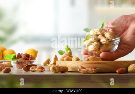 Mano con un recipiente pieno di arachidi su un tavolo di legno e cucina sullo sfondo. Vista frontale. Foto Stock