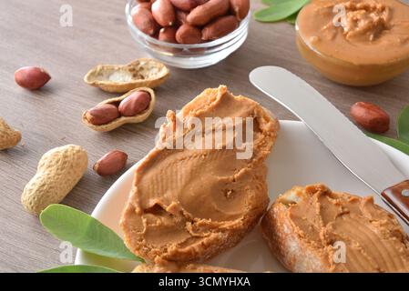 Dettaglio di ​​bread appena tagliato con burro di arachidi su un piatto bianco su un tavolo da cucina in legno marrone con prodotti e frutta. Vista dall'alto. Foto Stock