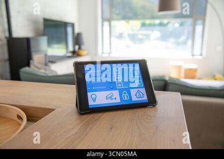 Tablet che mostra un'interfaccia di controllo intelligente per la casa, seduto su un tavolo in legno nel soggiorno con vista sul giardino. Tecnologia, automazione, interni, arredamento, contempo Foto Stock