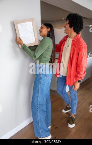 Coppia afroamericana e indiana appesa a quadri con cornice in legno su pareti bianche nell'area cucina. Opere d'arte, interni, arredamento per la casa, moderno, collaborazione, Foto Stock