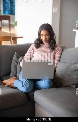 Donna afroamericana seduta a gambe incrociate su un divano grigio nel soggiorno utilizzando un computer portatile color argento. Intimità, produttività, moderno, comfort, tecnologia, casa Foto Stock