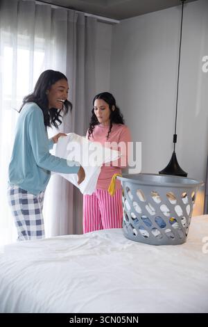 Diverse amiche, lavanderia pieghevole in camera da letto con cestino grigio e piumone bianco. Donne, amicizia, domestico, moderno, accogliente, tempo libero, vita Foto Stock