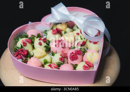 Una confezione regalo rosa a forma di cuore piena di fragole in cioccolato bianco e rosa, decorata con lamponi secchi e foglie verdi. Dessert elegante Foto Stock